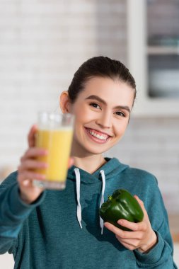 happy woman holding glass of smoothie and bell pepper while looking at camera on blurred foreground clipart