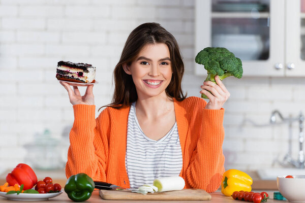 cheerful woman choosing between cake and fresh broccoli while smiling at camera
