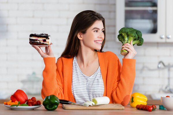 cheerful woman holding cake and fresh broccoli near vegetables in kitchen