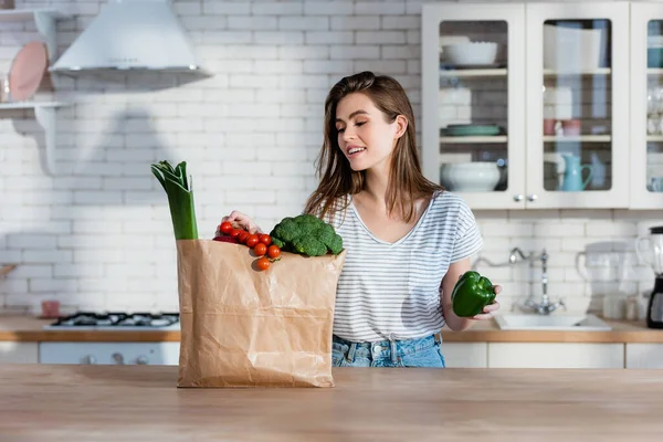 smiling woman holding bell pepper near paper bag with vegetables on kitchen counter