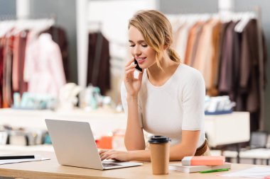 Smiling seller talking on smartphone and using laptop near coffee to go in showroom  clipart