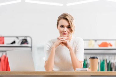 Dizüstü bilgisayarın, defterin ve kahvenin yanındaki kameraya bakan genç satıcı galeriye gidecek. 