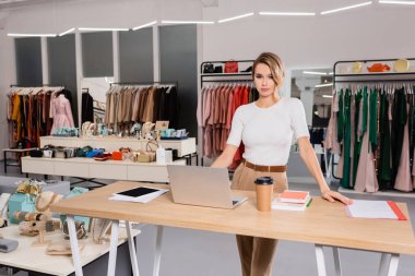 Galeride dizüstü bilgisayarın, defterin ve aksesuarların yanında duran satıcı. 
