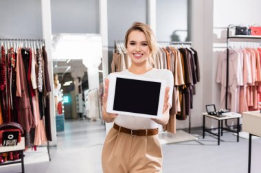 Neşeli satıcı sergi salonundaki bulanık önplanda boş ekranlı dijital tablet tutuyor 