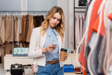 Akıllı telefon kullanan ve galeride kıyafetlerin yanında kağıt bardak tutan sarışın bir kadın. 