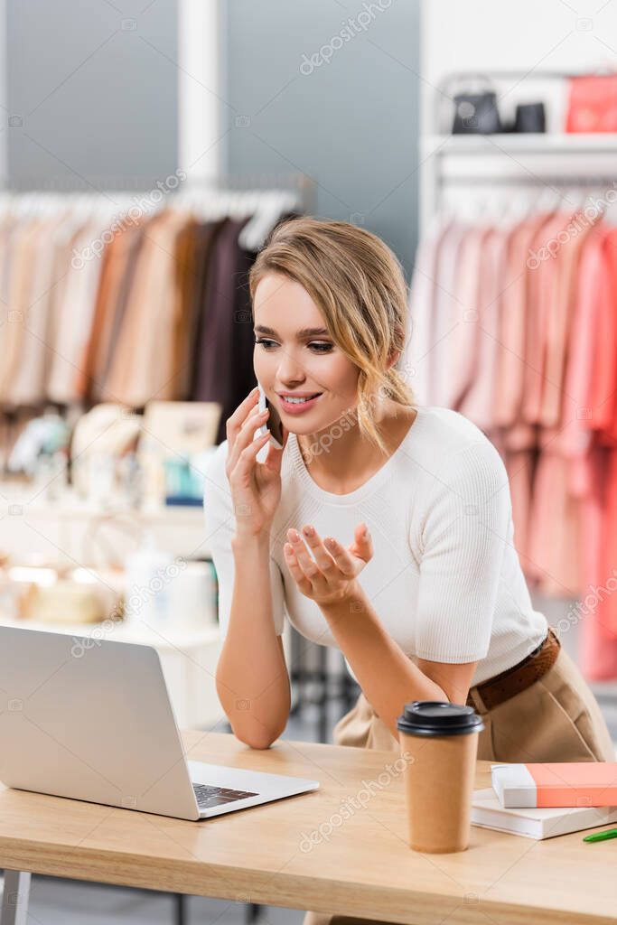 Young seller accepting order on cellphone near laptop and takeaway drink in showroom 