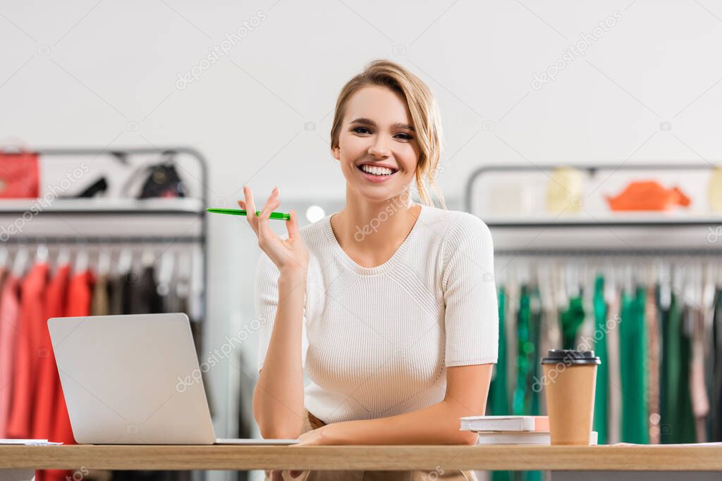 Positive seller holding pen near laptop and coffee to go in showroom 