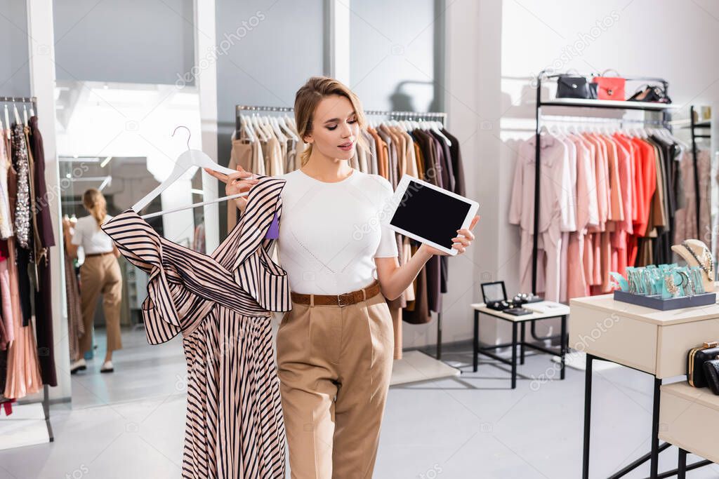 Pretty seller looking at digital tablet with blank screen while holding dress in showroom 