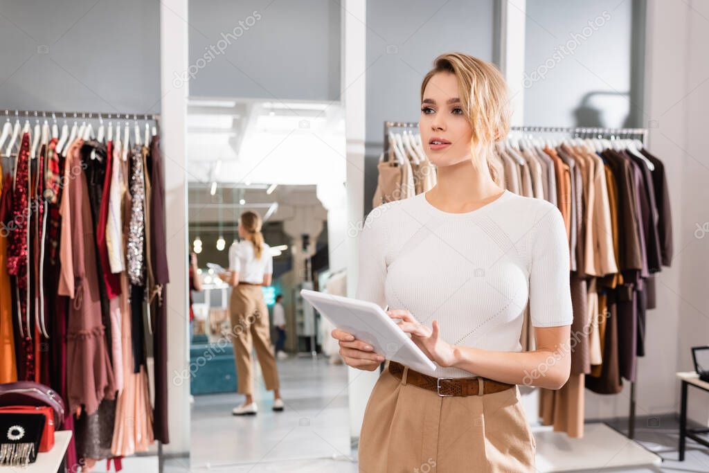 Blonde seller using digital tablet while working in showroom with clothes 