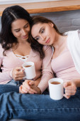 happy girlfriends holding cups of tea while sitting on sofa in living room