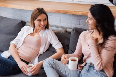 Mutlu kız arkadaşlar ellerinde çay fincanlarıyla kanepede oturup birbirlerine bakıyorlar.