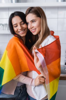 cheerful lesbian couple holding lgbt flag while hugging at home