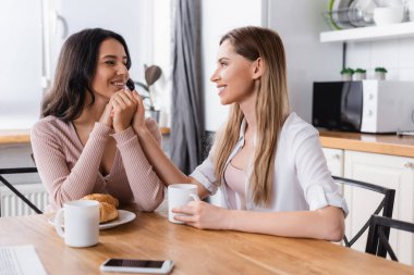 happy lesbian couple holding hands near breakfast and smartphone on kitchen table 