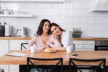 happy lesbian couple hugging near breakfast and smartphone on kitchen table 