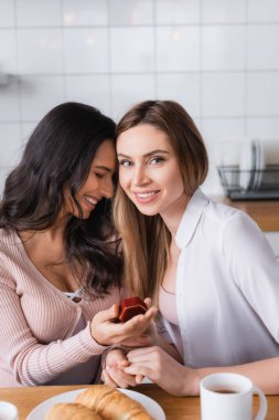 happy woman holding jewelry box with wedding ring near girlfriend in kitchen 