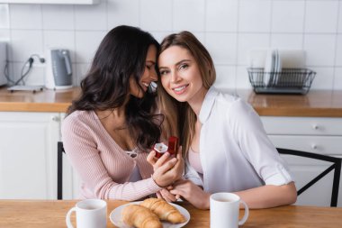 smiling woman holding jewelry box with wedding ring near girlfriend and breakfast in kitchen 