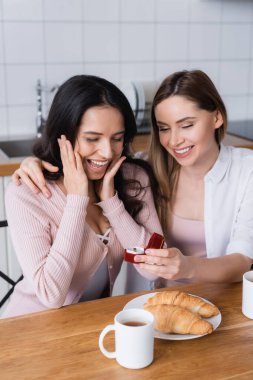 smiling woman holding jewelry box with wedding ring near excited girlfriend and breakfast in kitchen 