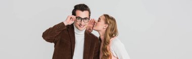 man in velvet blazer adjusting eyeglasses while girlfriend whispering in his ear isolated on grey, banner clipart