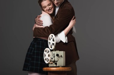 happy couple embracing near vintage film projector isolated on black clipart