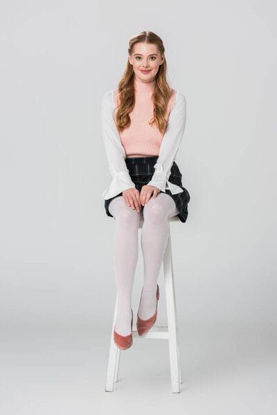 pretty, old fashioned woman smiling at camera while sitting on high stool on grey