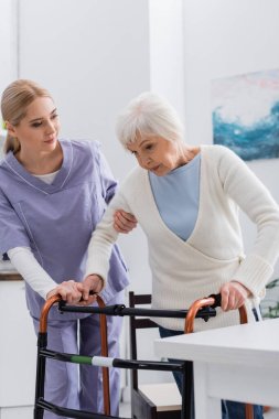 young social worker helping elderly woman walking with medical walkers at home clipart