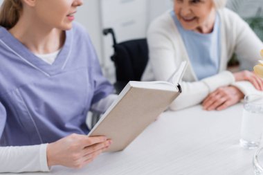 Genç hemşirenin yaşlı kadınlara kitap okuması, bulanık arka plan.