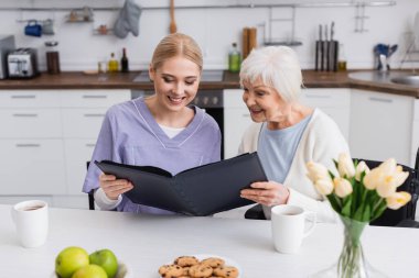 pleased senior woman and nurse looking at photo album in kitchen clipart
