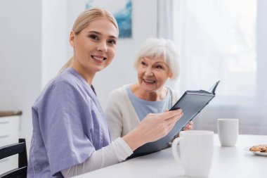 happy social worker looking at camera while holding photo album near aged woman clipart