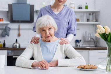 young nurse touching shoulders of elderly woman looking at camera near jigsaw puzzle clipart