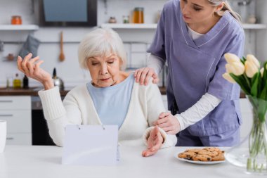 young nurse touching shoulder of senior woman showing shrug gesture near calendar clipart