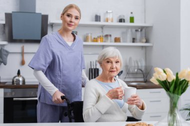 young nurse looking at camera near handicapped senior woman with cup of tea in kitchen clipart