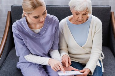 young nurse and senior woman, suffering from memory loss, pointing at calendar while sitting on couch clipart