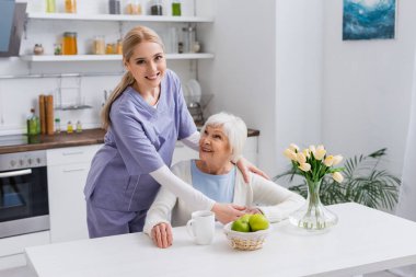 Mutfakta mutlu yaşlı bir kadını kucaklarken kameraya gülümseyen genç hemşire.