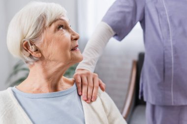 İşitme cihazı olan yaşlı bir kadın omzuna dokunan sosyal hizmet görevlisine bakıyor.