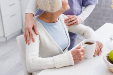 partial view of nurse hugging shoulder of aged woman sitting near cup of tea clipart
