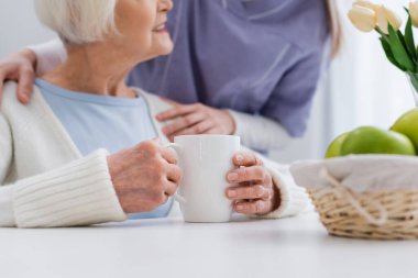 Çay fincanının yanında gülümseyen yaşlı bir kadının omzunu kucaklayan hemşire manzarası.
