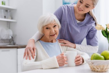 happy aged woman smiling at camera while young nurse hugging her shoulders in kitchen clipart