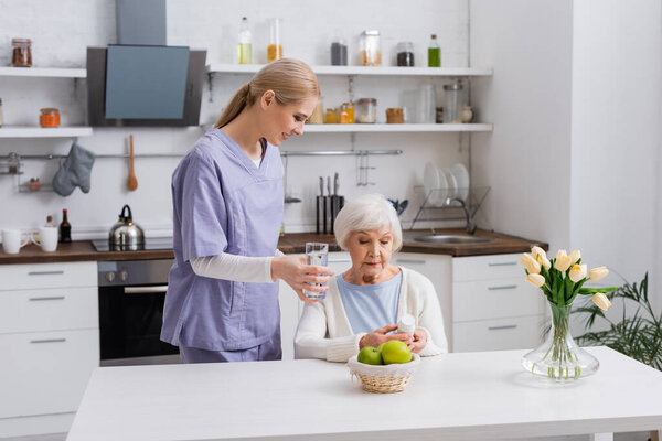 young nurse giving glass of water to elderly woman holding pills container in kitchen