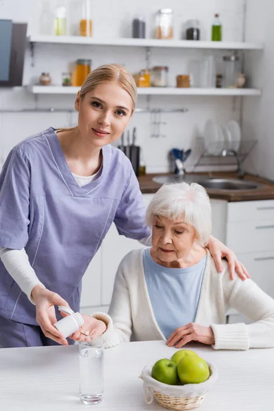 Ung Sjuksköterska Tittar Kameran Medan Ger Piller Till Äldre Kvinna — Stockfoto