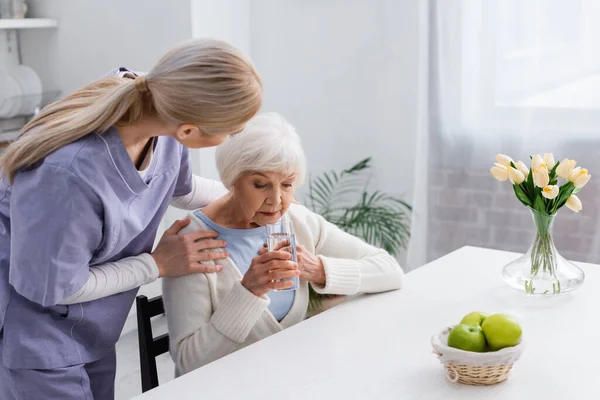 Joven Enfermera Abrazando Hombros Anciana Sosteniendo Vaso Agua Mientras Está — Foto de Stock