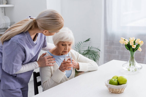 young nurse embracing shoulders of elderly woman holding glass of water while sitting near table