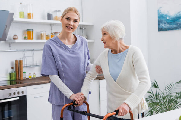 young nurse smiling at camera while supporting aged woman near medical walkers