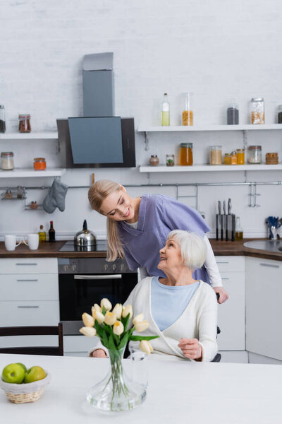 young social worker looking at happy elderly woman sitting in kitchen