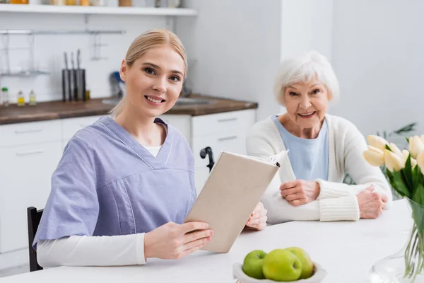 Enfermera Sonriente Con Libro Mirando Cámara Cerca Feliz Anciana Cocina —  Fotos de Stock