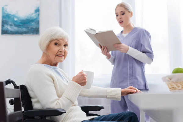看護師の近くのカメラを見ているお茶を飲んでいる障害のある高齢者の女性背景のぼやけた本 — ストック写真