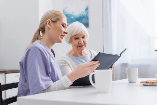 Smiling Nurse Happy Elderly Woman Looking Photo Album Cups Tea — Stock Photo, Image