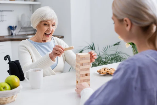 Podekscytowana Seniorka Gra Drewnianą Wieżę Pracownikiem Socjalnym Niewyraźnym Pierwszym Planie — Zdjęcie stockowe