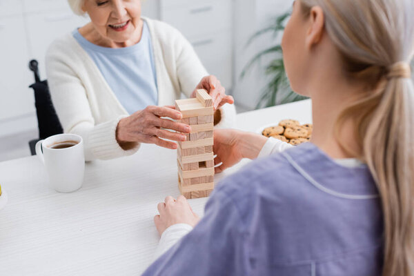 обрезанный вид радостной пожилой женщины, играющей в деревянные блоки игры с медсестрой на размытом переднем плане