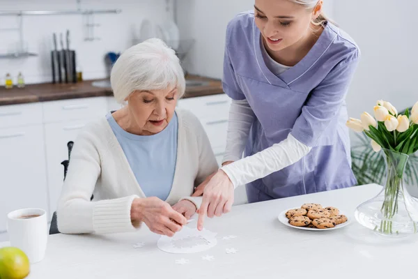 Joven Enfermera Señalando Rompecabezas Cerca Anciana Mujer Cocina —  Fotos de Stock
