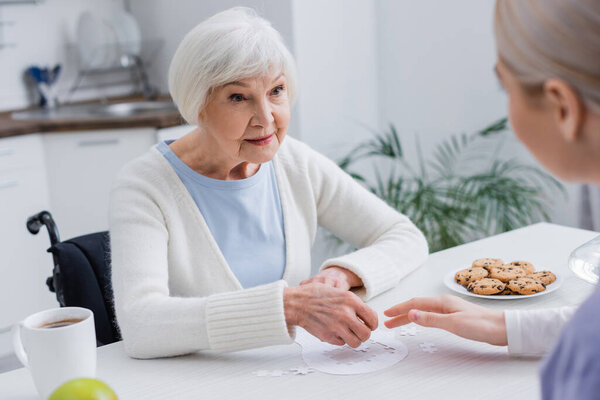 elderly woman playing jigsaw puzzle with social worker on blurred foreground